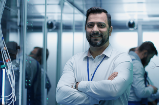 Male IT Professional Standing with Arms Crossed in Server Room with others in the background